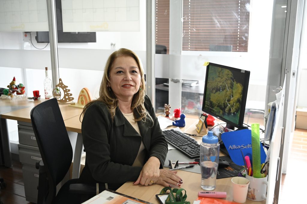 Retrato de una mujer profesional sonriendo en su oficina, sentada frente a un escritorio con computadora y ventanales transparentes de fondo.