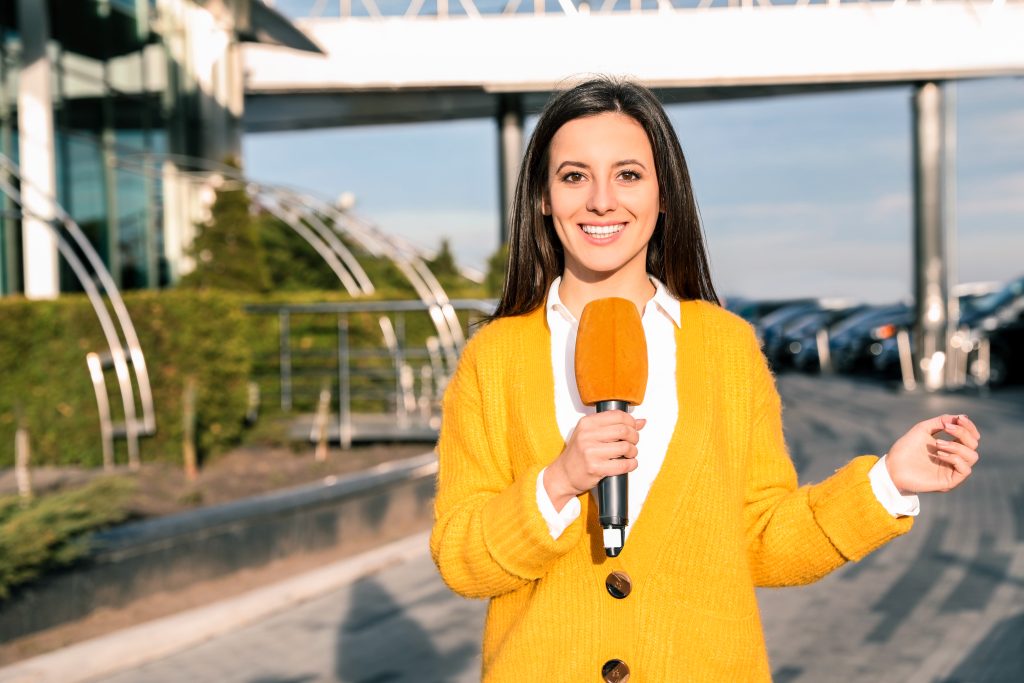 Periodista sonriente con micrófono naranja reportando en exteriores frente a edificio corporativo.