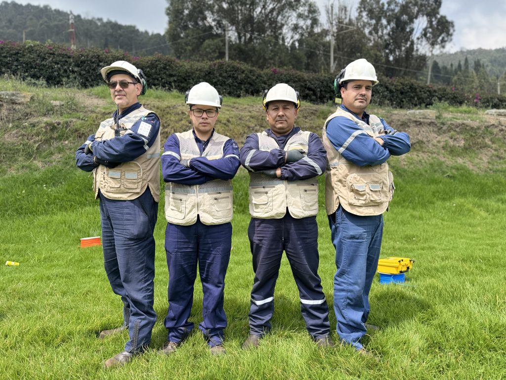 Cuatro operarios con cascos blancos, chalecos beige y monos azules posan de brazos cruzados en un campo verde.